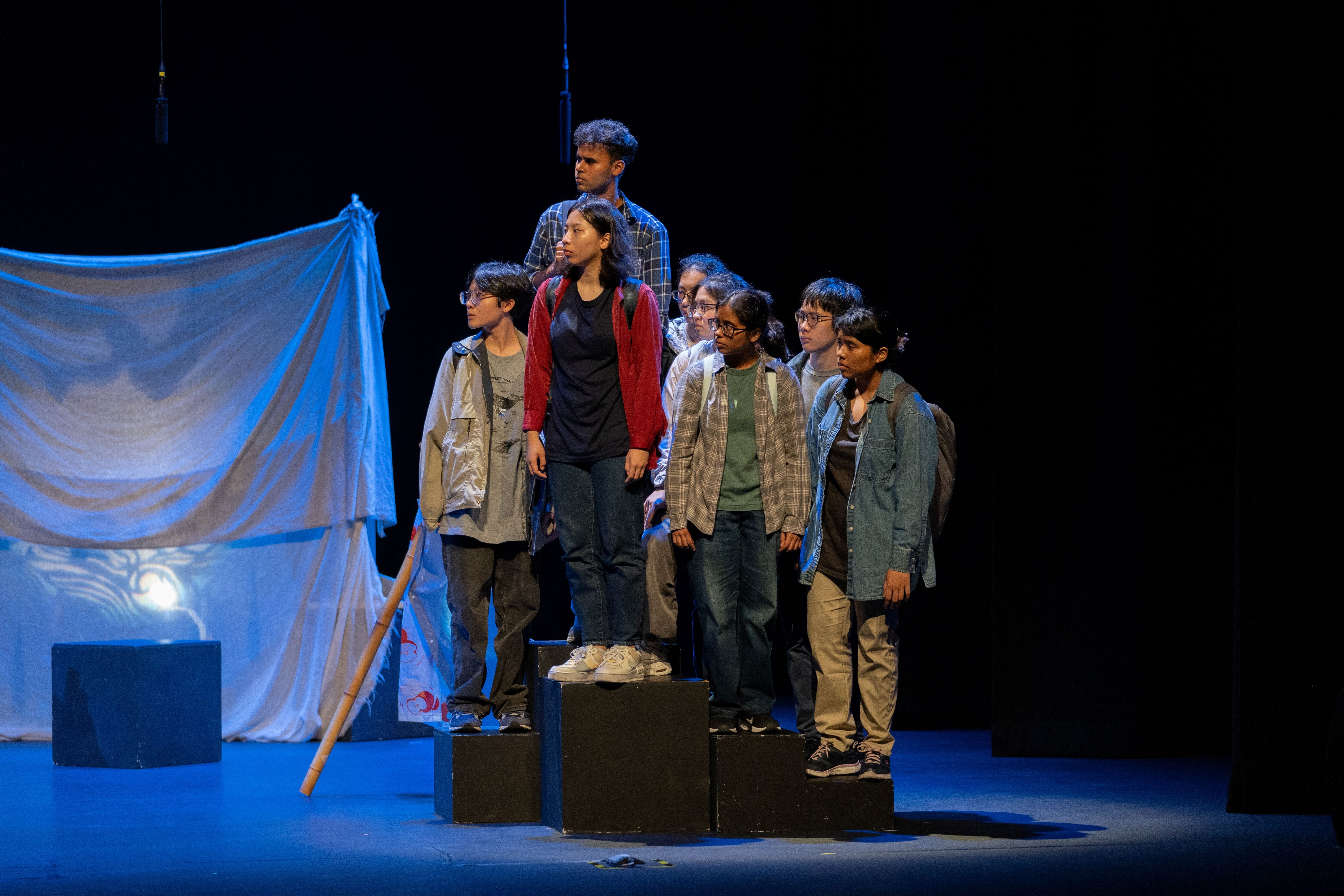 Eight student performers stand together on raised black platforms under cool blue stage lighting, with a large draped white fabric backdrop behind them.